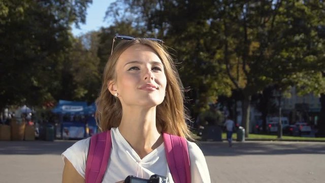Beautiful caucasian fair hair tourist girl, with a pink backpack, is taking a photo in the street, sunny day, slow motion