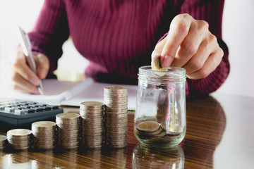Woman process and drop coin into the glass with stack coins and calculator. Woman working on table.