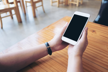 Mockup image of hands holding white mobile phone with blank black screen on wooden table in cafe