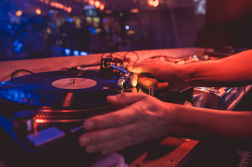 Hands of a DJ putting a vinyl record on a record player in a music club in the rays of blue and red...