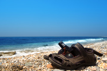 Fototapeta na wymiar Pair of old sandals and sunglasses on the pebble beach of the Mediterranean Sea