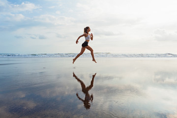 Barefoot sporty girl with slim body running along sea surf by water pool to keep fit and health. Beach background with blue sky. Woman fitness, jogging sports activity on summer family vacation.