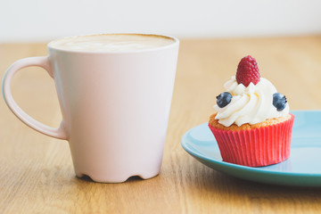 Small cupcake with raspberry and blueberry on the plate, cup of hot cappuccino