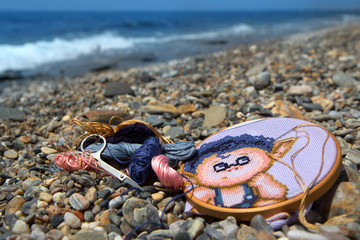 Cross stitch embroidery, multicolored threads, needle, scissors and hoop on the pebble beach of the Mediterranean Sea