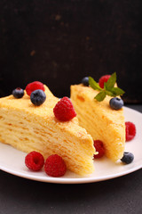 Napoleon cake decorated with raspberries, blueberries and mint on a white plate on a black background