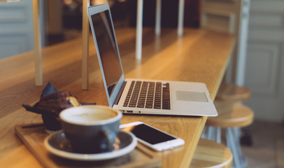 A close up view of mug of coffee placed behind a laptop with a blank screen on the blurred background. An opened portable computer and smartphone is placed with a cup on a wooden table in a cafe. - obrazy, fototapety, plakaty