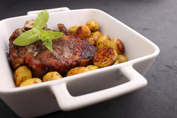 Steak on a bone baked in white dish with vegetables: potatoes, garlic, onions. Black background