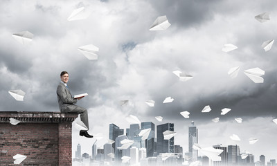 Man on roof edge reading book and cityscape at background