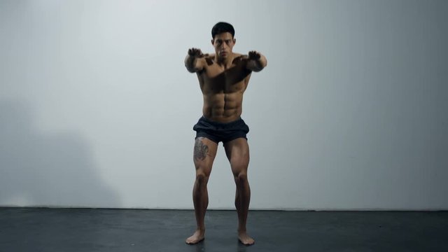 A chiseled fitness model performs squats in front of a grey wall. Long shot.