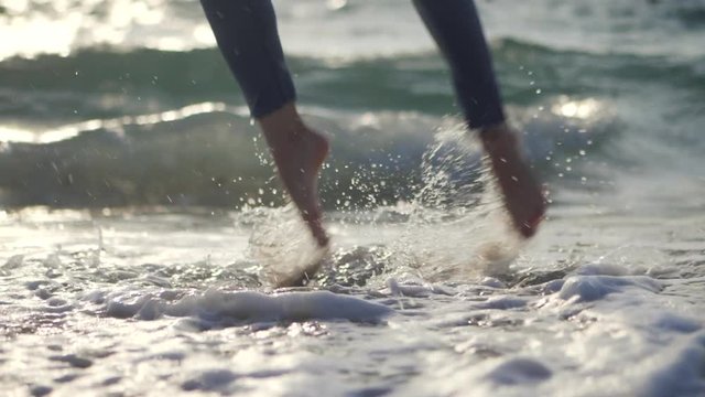 Close up of a ballet dancer's feet as she practices barefoot in sea or ocean water. Ballerina demonstrates classic ballet pas and jumping in casual style - demim. Slow motion.