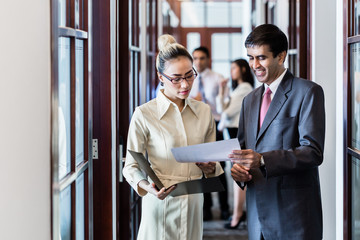 Indian business executive man and Indonesian secretary standing in office hallway discussing papers