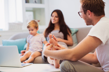 Close-up young man doing foot massage his wife who sitting on the sofa with little son and looking laptop