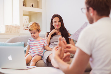 The young man doing foot massage his angry wife who sitting on the sofa and looking at documents near little son who watching laptop