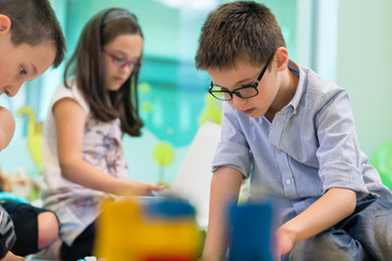 Cute girl wearing eyeglasses while playing with concentration a creative game next to her colleague in the classroom of a modern kindergarten 