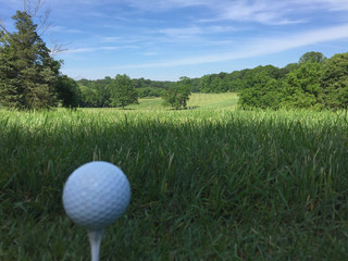 White golf ball sits on a tee ready to be hit by club down the fairway of a beautiful generic golf course on a sunny day