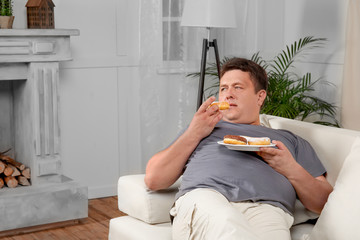 Overweight young man eating sweets on sofa at home