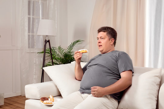 Overweight young man eating sweets on sofa at home