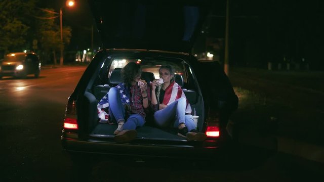 Beautiful Girls Sitting In The Open Trunk Of The Car, Talking And Eating Burgers During The Night In The City. Slowmotion Shot