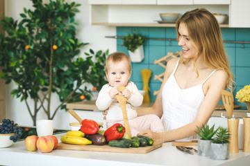 Young beautiful mother is cooking and playing with her baby daughter in a modern kitchen setting. Healthy food concept.