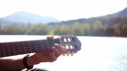 Close up of hand guy plays on a guitar sitting by mountain river on sunny day