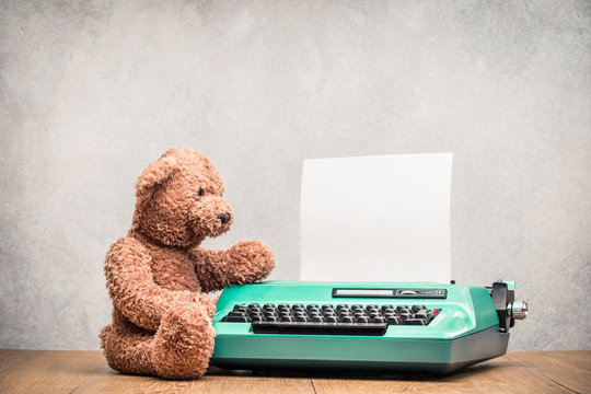 Retro Teddy Bear toy typing on old classic mint green typewriter with paper sheet on wooden desk front concrete wall background. Vintage instagram style filtered photo