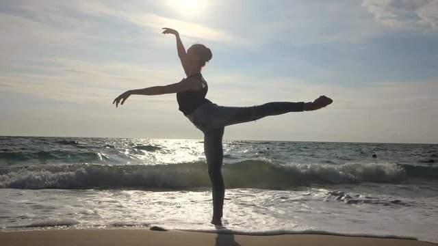 Silhouette of young woman in casual style - denim and black top doing ballet at the beach. Attractive ballerina practices in stretching on sandy coastline in autumn. Slow motion. Gimbal shot
