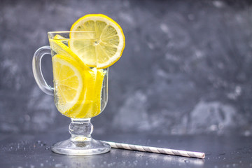 Homemade lemonade in a glass with a handle on a gray concrete background. Water with slices of lemon on a dark background.