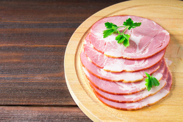 Sliced ham with fresh green lettuce leaves on a round cutting board. Meat products on a brown wooden table.