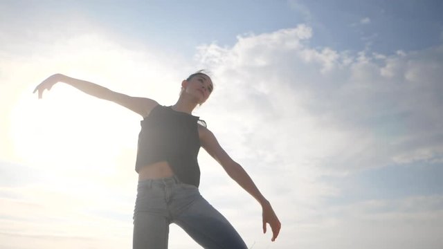 Beautiful scene of dancing ballerina in black top and jeans on beach near ocean or sea in morning. Beautiful brunette woman practicing hands exercises. Flare. Gimbal shot. Slow motion.