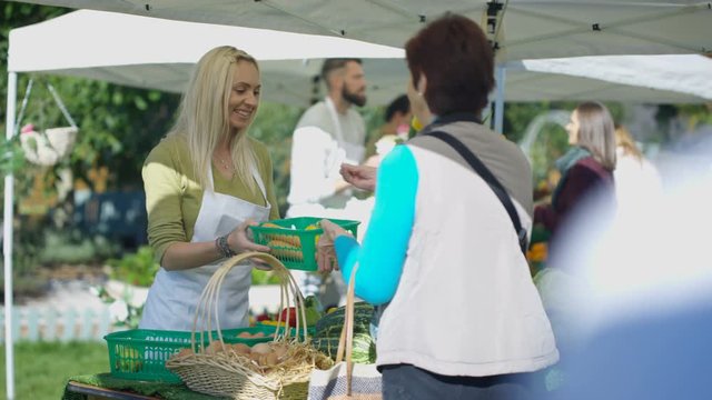  Cheerful woman selling fresh fruit & veg to customers at farmers market