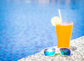 Sunglasses with orange juice and a piece of lemon at the side of swimming pool. Vacation, beach, summer travel concept