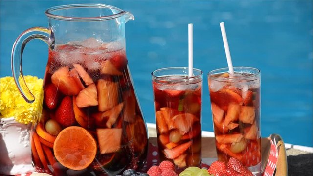 Summer pool party - sangria pitcher with glasses, fresh fruit and French macaroons on a table near a turquoise swimming pool in a garden setting

