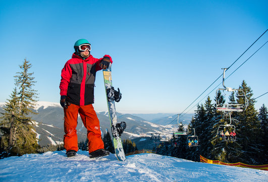 Full length portrait of a snowboarder wearing helmet skiing mask and colorful winter snowboarding clothing standing with his snowboard looking away in the evening copyspace active lifestyle sports