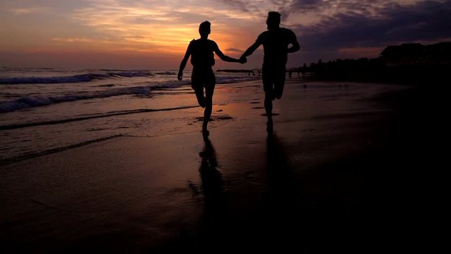 Happy couple holding each other hands and running on the beach, slow motion shot at 240fps, steadycam shot

