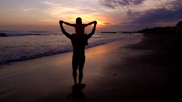 Father gives a son a piggyback on the beach, slow motion shot at 240fps, steadycam shot
