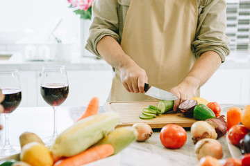 The man is cutting the cucumber in the kitchen in his house to prepare salad along with eating dinner.