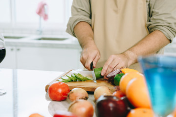 The man is cutting the cucumber in the kitchen in his house to prepare salad along with eating dinner.