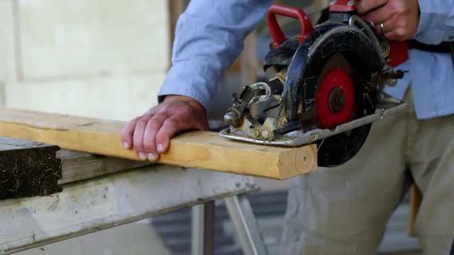 Detail of a contractor using a hand-held electric circular saw to cut through a piece of wood inside a house under construction. Slow motion 4K recorded at 60fps with slider move.