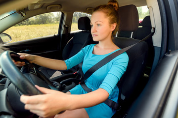 Obraz na płótnie Canvas Young casual woman in blue dress driving a car, side view. Beautiful young girl at the wheel of car with black interior looking at the road
