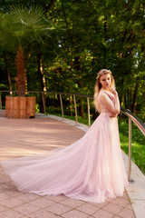 Fototapeta premium Portrait of a beautiful bride blond girl in pink lace dress ,hair decoration, handmade. Tenderness. Standing in the midst of a dark forest green in the rays of the setting sun. Shows a long dress with