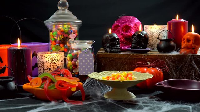4k Happy Halloween trick or treat party table with bowls and apothecary jars of candy with skull candles against a black background, scooping candy into bowls.