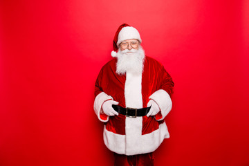 Have a holly jolly x mas! Gifts for kids! Festive seosonal  occasion. Funny Santa Claus wearing red traditional outfit, adjusting big black belt, standing isolated on red background, copy space