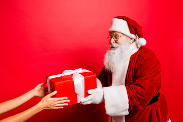 Holly jolly x mas! Festive seosonal occasion. Funny Saint Nicholas in red traditional fur coat presenting a wrapped gift with white tape, cropped female hands accepting it, isolated on red background