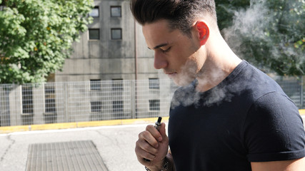 Handsome man standing in city street and vaping with electronic cigarette, then smiling to camera