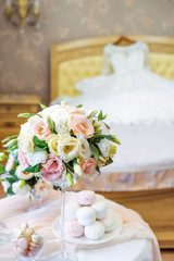Table with bride's details: bouquet of eustoma flowers, jewelery, perfumes and marshmallow and macaroon sweets with blurred Wedding dress lying on the bed on the background. Selective focus