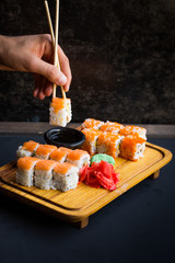 Salmon sushi rolls, sashimi and wasabi on traditional wooden serving tray, Japanese Chinese  eastern cultures asian food. Male hand with wooden bamboo chopsticks holding a sushi over the soy sauce 