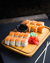Salmon sushi rolls, sashimi and wasabi on traditional wooden serving tray, Japanese Chinese eastern cultures asian food. Wooden bamboo chopsticks by side on dark table and black background 
