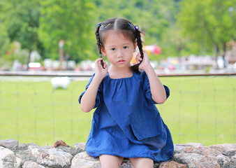 Little asian girl sitting on the stone wall and touching her two tied ponytails hairs at green public garden.