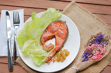 Fried salmon steak with vegetables and knife - fork on a white plate against wood plank background. Decoration with dried flower.