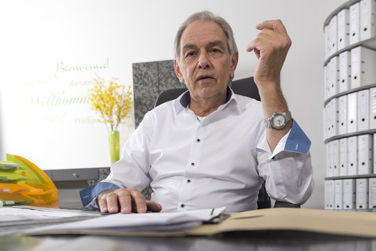 &Auml;lterer Mann mit wei&szlig;em Hemd  sitzt an einem Schreibtisch und bl&auml;ttert in einer Dokumenten Akte
